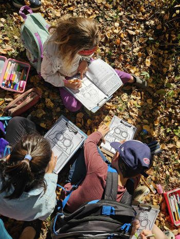 Trois enfants assis dans les feuilles, avec crayons et cahiers d'exercices
